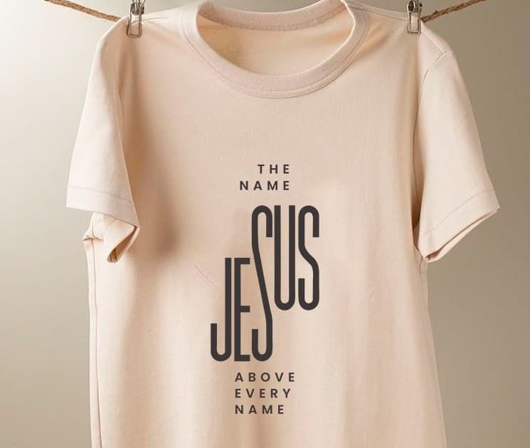 A cream ADULLAM T-shirt printed with “The Name Jesus Above Every Name”, hanging from a line.